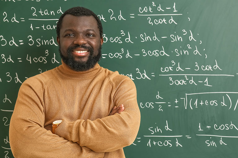Man wearing a yellow sweater crossing arms and standing in front of equations on a blackboard