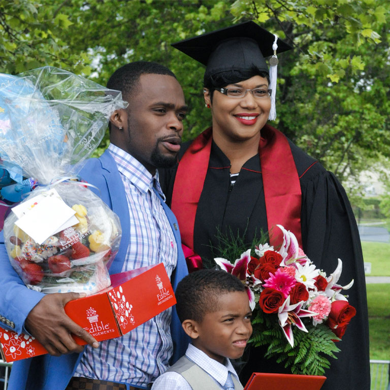 A proud family takes a picture with a female graduate. The man is loaded with Commencement gifts and he and the little boy also pictured are wearing summer suits.