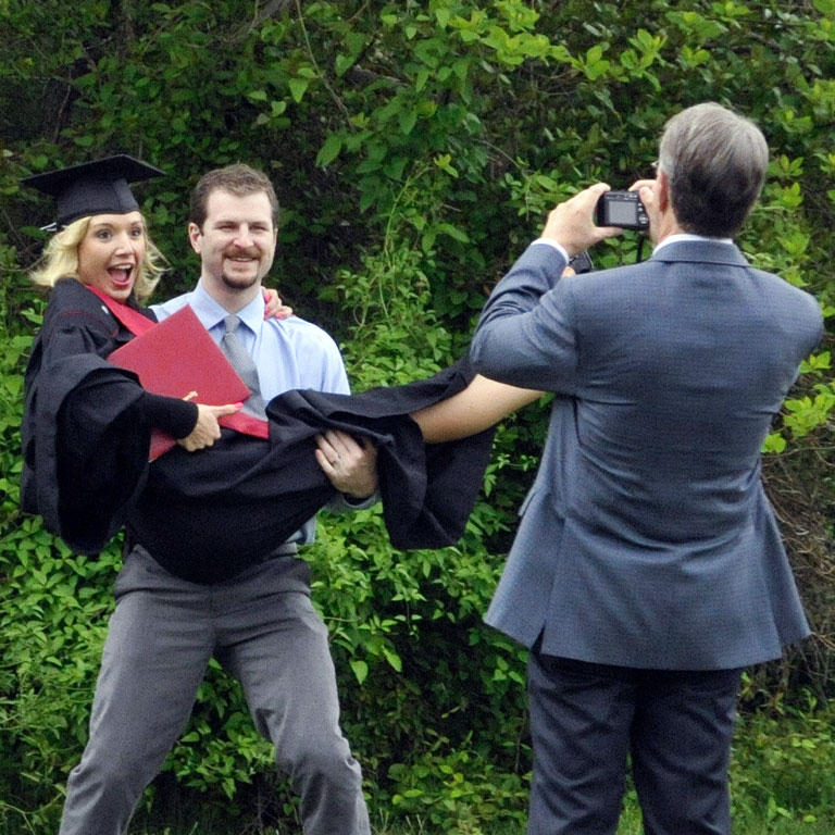 A young man in suit and tie picks up a laughing female graduate as their family takes pictures