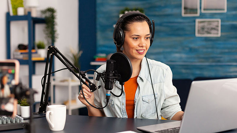 Woman wearing headphones, looking at a laptop, and speaking into a microphone while being recorded by a phone
