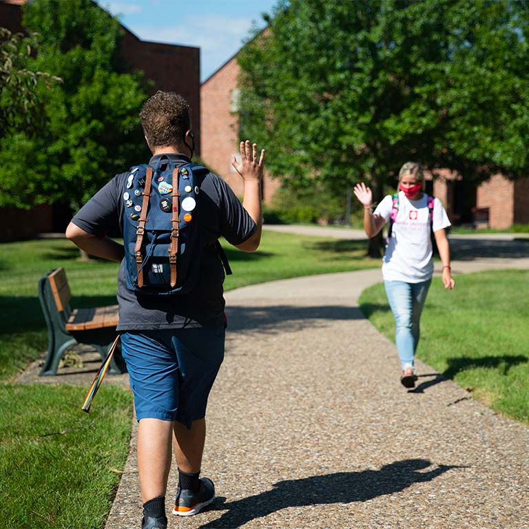 Male and female students waving to each other as they pass on the sidewalk. 